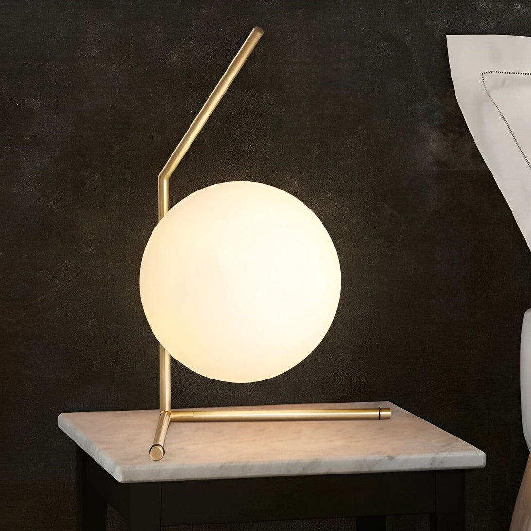 Une lampe de table en métal doré avec un abat-jour en verre opalin blanc. La lampe est posée sur une table de chevet en marbre blanc. CA-2757427814,CA-2757537830,