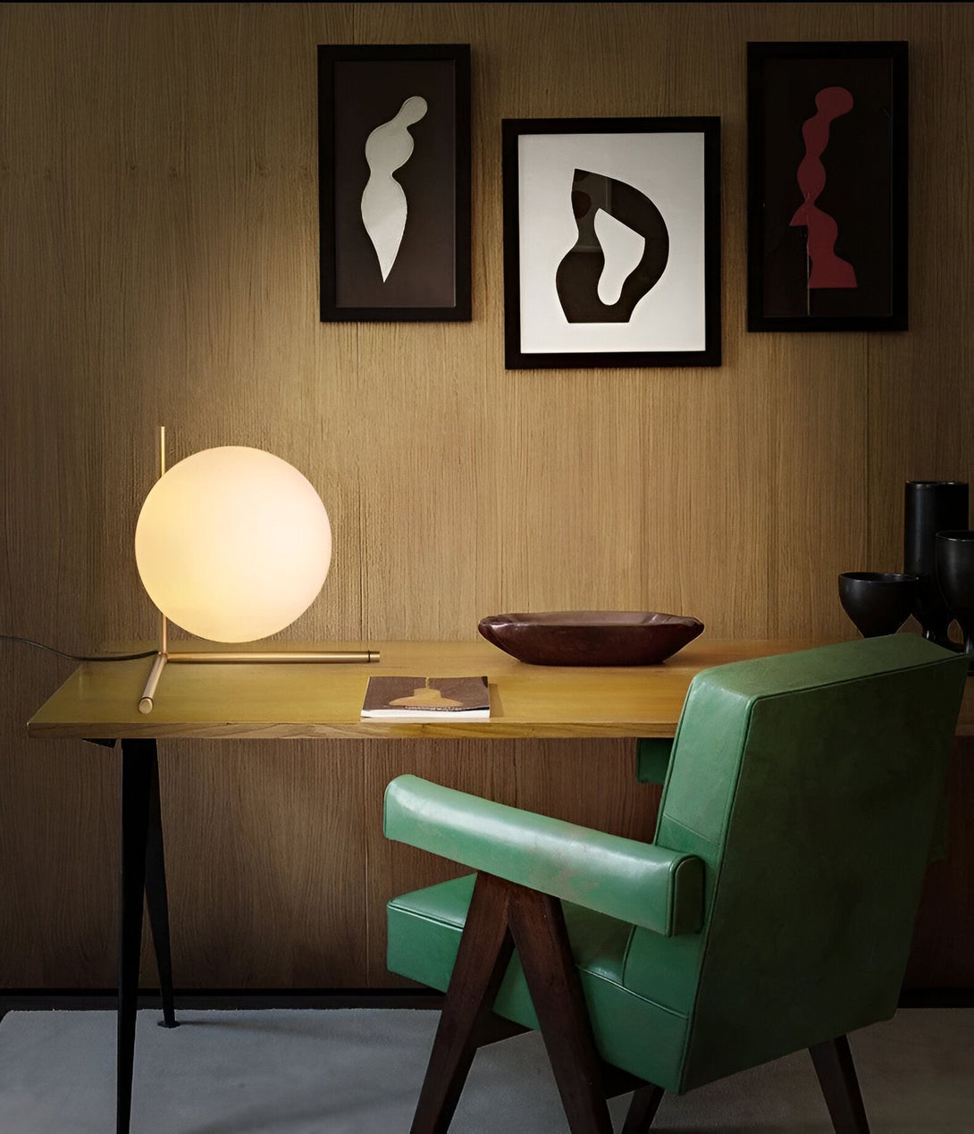 Une lampe de table en verre soufflé opalin avec une base en métal doré. La lampe est posée sur un bureau en bois avec un livre et un bol en bois. Derrière le bureau, il y a un fauteuil vert et trois cadres noirs accrochés au mur. CA-2757427814,CA-2757537830,
