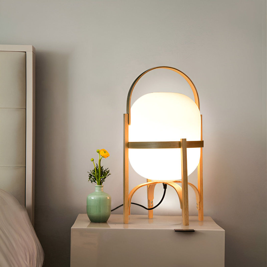 Une lampe de chevet en bois avec une anse en métal et un abat-jour en verre blanc. À côté de la lampe, sur une table de chevet, se trouve un vase vert contenant une fleur jaune. BU-5057562819,