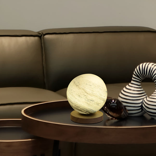 Une lampe sphérique en verre dépoli est posée sur une table basse en bois. À côté d'elle se trouve une sculpture en métal noir et blanc. Derrière la table basse, se trouve un canapé en cuir marron. OR-7630686498,OR-0614193508,