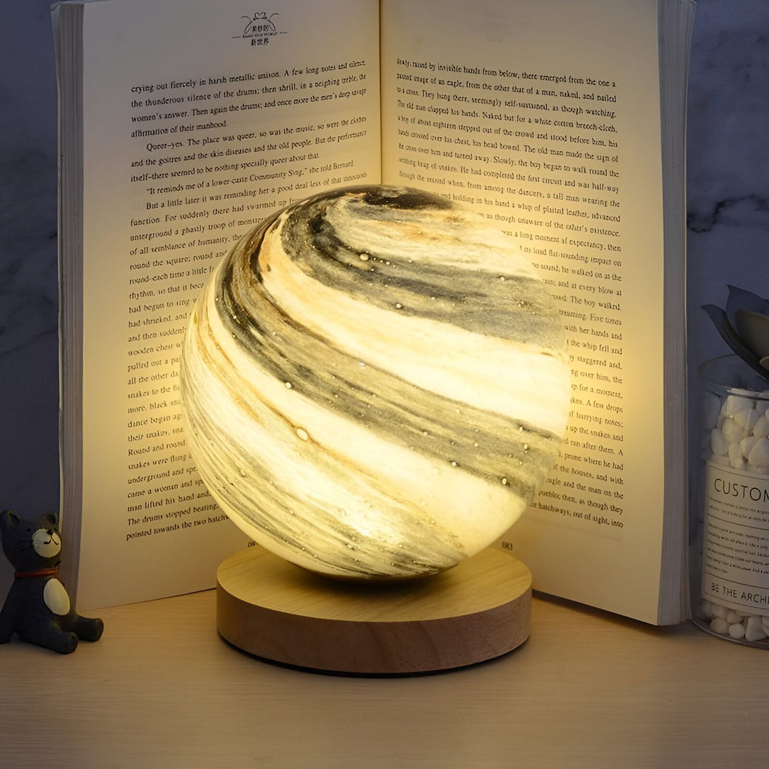 Une image d'une lampe en verre soufflé en forme de planète. La lampe est posée sur un socle en bois. Elle est allumée et diffuse une lumière douce. OR-7630686498,OR-0614193508,