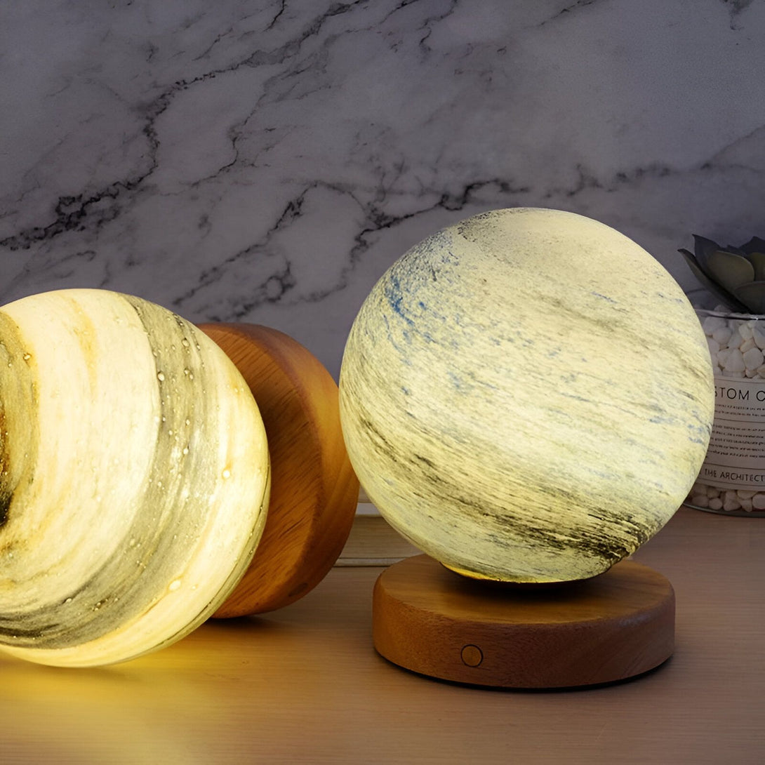 Une lampe en verre soufflé en forme de planète. La lampe est posée sur un socle en bois. La lampe est allumée et émet une lumière douce. OR-7630686498,OR-0614193508,