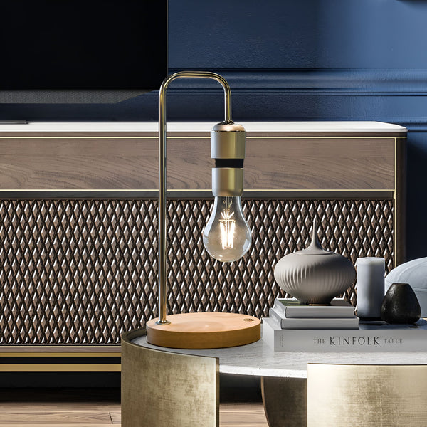 Une lampe à poser design en métal doré avec une base en bois clair. L'ampoule est maintenue en lévitation par un champ magnétique. À côté se trouve une table basse avec un livre, un vase et des bougies. LE-5812217572,