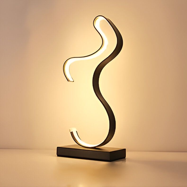 Une lampe de table noire en forme de S avec une base rectangulaire noire. La lampe est allumée et émet une lumière blanche chaude. EV-1596845252,EV-6746318540,EV-1161783687,