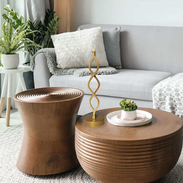 Une lampe dorée en forme de spirale est posée sur une table basse en bois. À côté d'elle, se trouve une plante verte dans un pot blanc. Derrière la table basse, il y a un canapé gris avec des coussins blancs et gris. IN-761638868X,IN-2865725197,IN-8287419824,IN-8037118681,