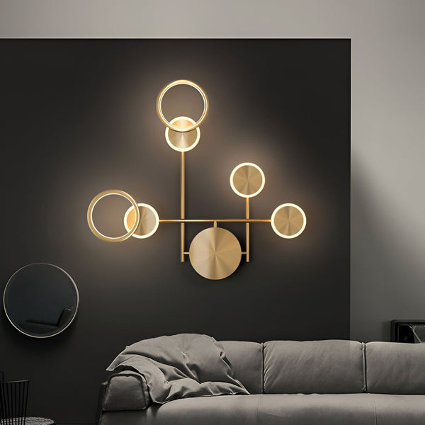 Une image d'une applique murale en métal doré avec cinq anneaux lumineux. L'applique est fixée sur un mur gris foncé et éclaire le mur d'une lumière chaude. DO-3701537453,DO-1406931985,DO-774364684X,DO-8119607104,