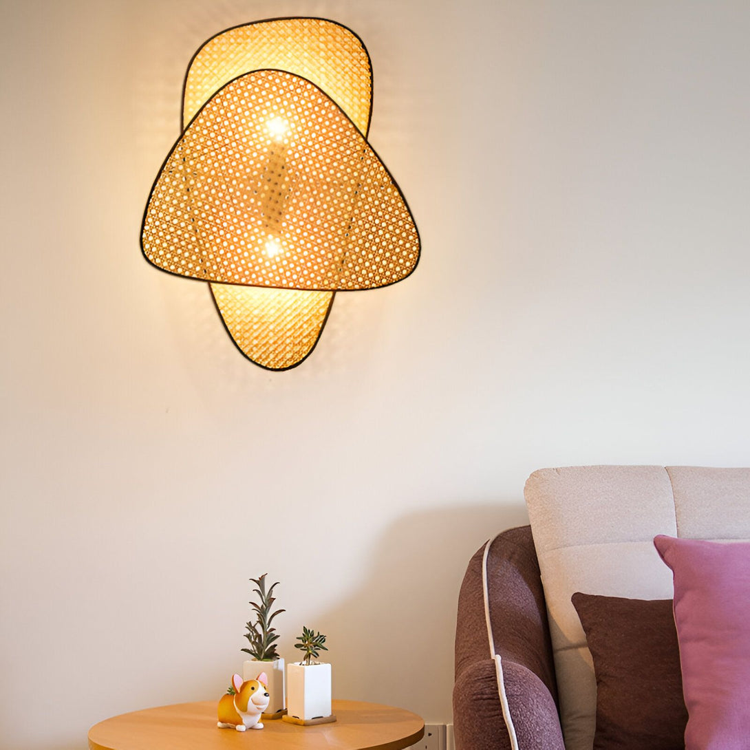 Une applique murale en osier tressé en forme de triangle avec une lumière chaude. À côté, se trouve un canapé marron avec des coussins blancs et roses. Sur la table basse, il y a un chien en céramique, une plante verte et une plante grasse. BA-0203883330,BA-3132460699,