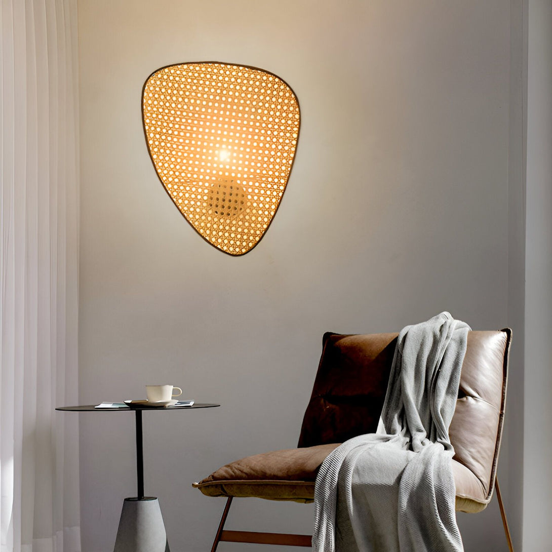 Une applique murale en forme de losange en osier avec une lumière allumée à l'intérieur. À côté, se trouve un fauteuil en cuir marron avec un plaid gris dessus. BA-0203883330,BA-3132460699,