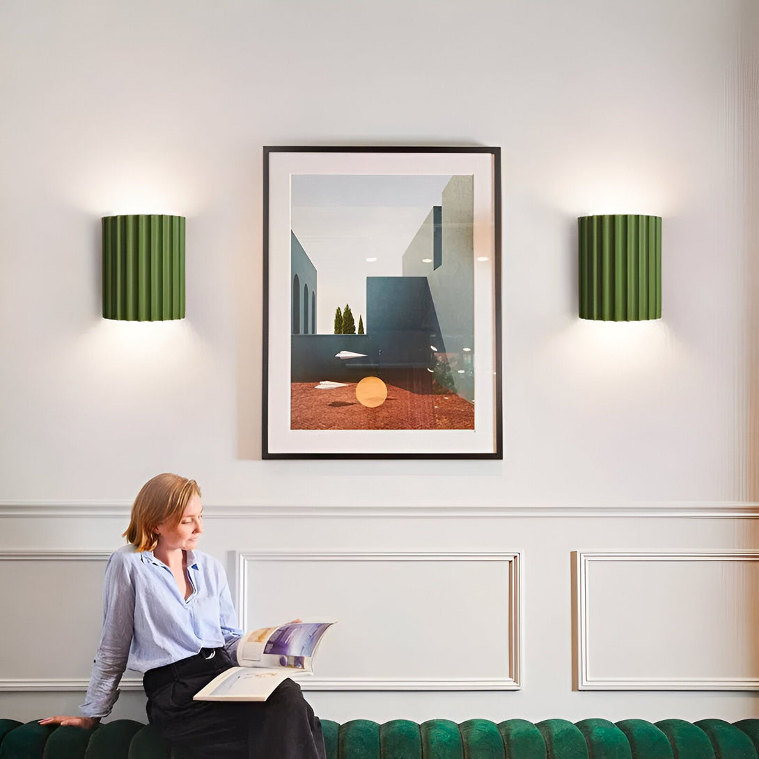 Une femme est assise sur un canapé vert et lit un magazine. Il y a deux appliques murales vertes de chaque côté d'un tableau représentant un paysage urbain. NE-721526775X,NE-8904744822,NE-2805852109,NE-679239677X,