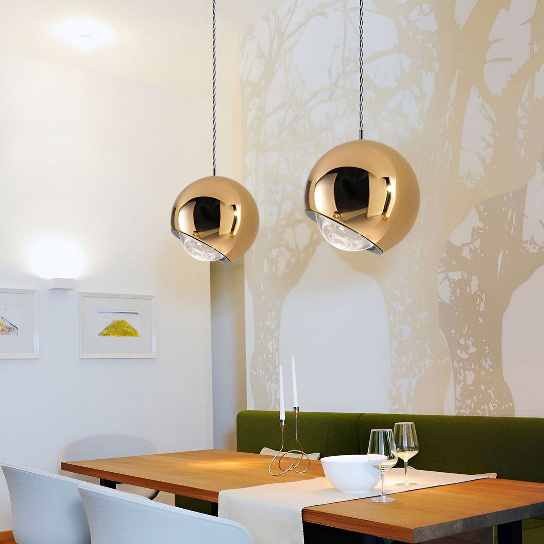 Une image de deux suspensions dorées en forme de sphère. Les suspensions sont accrochées au plafond par un fil noir et sont allumées. Il y a une table en bois avec une nappe blanche et des verres à vin dessus. Derrière la table, il y a un mur avec un arbre peint dessus. OB-1416333185,OB-1568639201,OB-983265761X,OB-4434955144,OB-3129829164,OB-0998241695,OB-6069405625,OB-5906347348,OB-9038855125,OB-7633987596,OB-8882831361,OB-4666004629,
