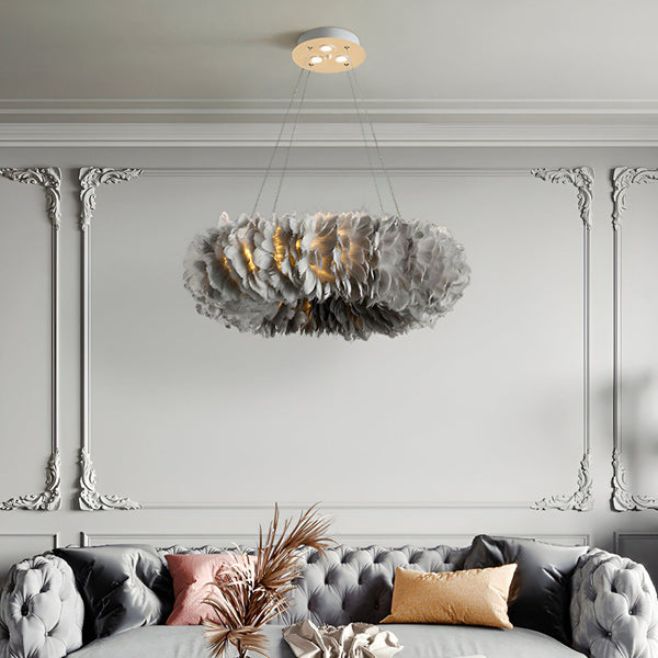 Une image d'un lustre en plumes d'autruche gris suspendu au plafond. Le lustre est rond et composé de plusieurs couches de plumes. Il est suspendu par trois chaînes et éclairé par des ampoules LED. Le lustre est placé dans un salon avec un canapé gris et des murs blancs.