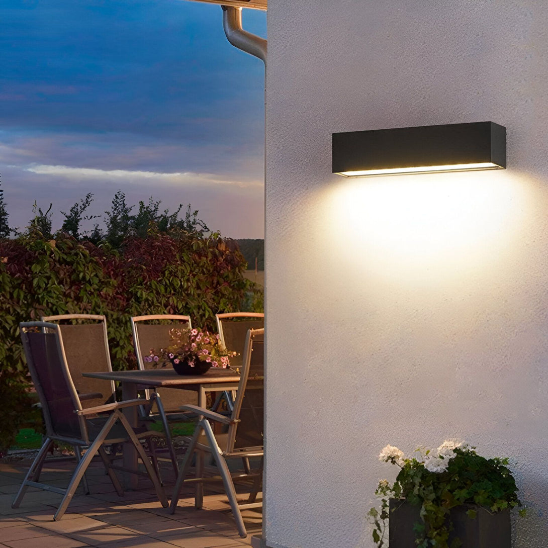 Une applique murale extérieure noire émet une lumière blanche chaude sur un mur extérieur. À côté de l'applique, se trouve une porte et des chaises de jardin. NA-033735863X,NA-9496728251,NA-8762483625,NA-6848322024,NA-1955103321,NA-7992054555,NA-9701687752,NA-4955698107,