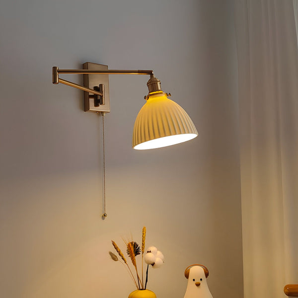 Une image d'une applique murale en laiton avec un abat-jour en céramique blanche. L'applique est montée sur un mur blanc et est allumée. Il y a un vase avec des fleurs séchées et un oiseau en céramique blanche sur une table à côté de l'applique. YG-3474575996,YG-1688524622,YG-6005096354,YG-629266189X,YG-0652226035,