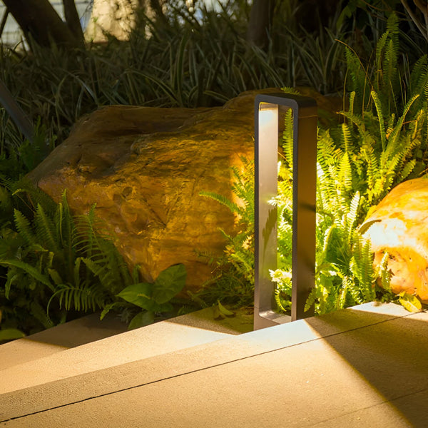 Une image d'une lampe de jardin en métal noir. La lampe est de forme rectangulaire et émet une lumière blanche. Elle est posée sur une dalle en bois, à côté d'un rocher et de plantes vertes. NE-755254144X,NE-2769874705,NE-0294437452,NE-1666104728,NE-5327672107,NE-988439248X,NE-8976410963,NE-906375275X,