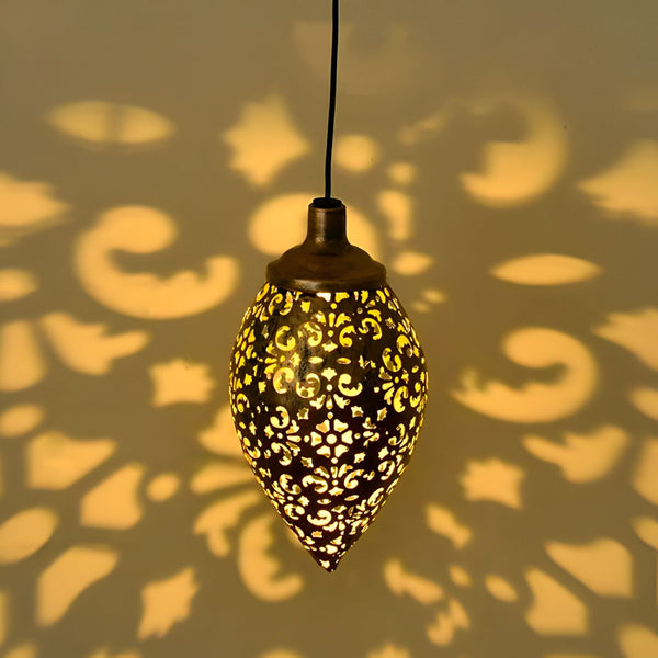 Une lanterne marocaine en métal ajouré de couleur bronze avec une lumière chaude à l'intérieur. La lanterne est suspendue par un fil noir et projette des ombres complexes sur le mur. Le produit est nommé Luminaire Extérieur Solaire Jardin. SU-0023998237,