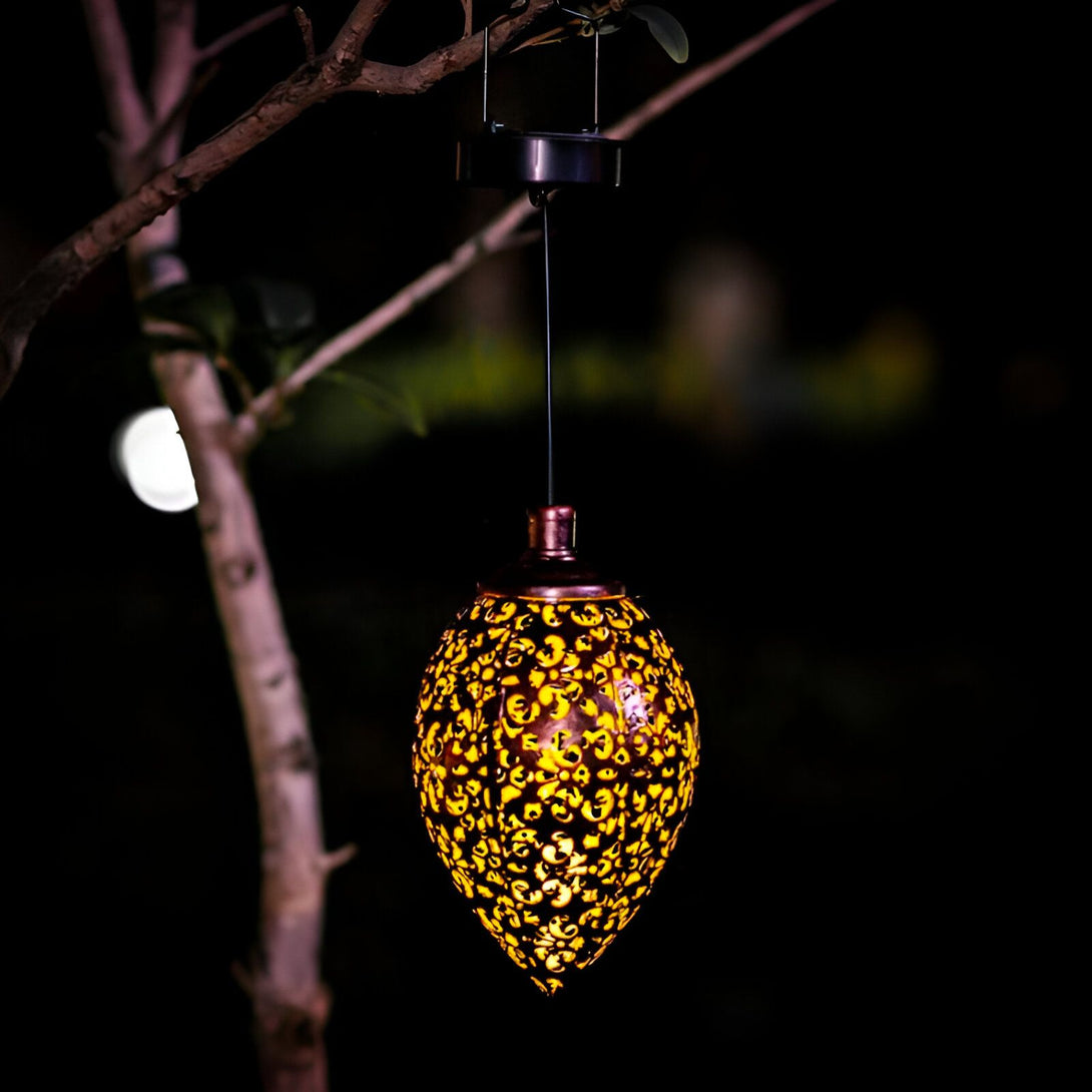 Une lanterne solaire d'extérieur en forme de poire est suspendue à une branche d'arbre. La lanterne est en métal et est de couleur cuivre. Elle est décorée de motifs floraux. La lanterne est éclairée par une lumière LED jaune. SU-0023998237,