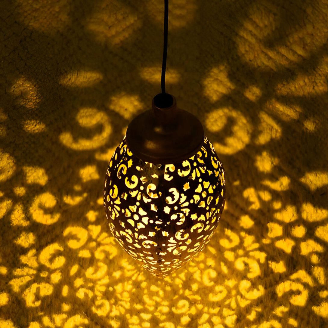 Une lanterne solaire d'extérieur en métal de couleur bronze avec un motif floral ajouré qui projette de belles ombres sur le sol lorsqu'elle est allumée. La lanterne est suspendue par une chaîne et est dotée d'un panneau solaire sur le dessus pour charger la batterie. SU-0023998237,