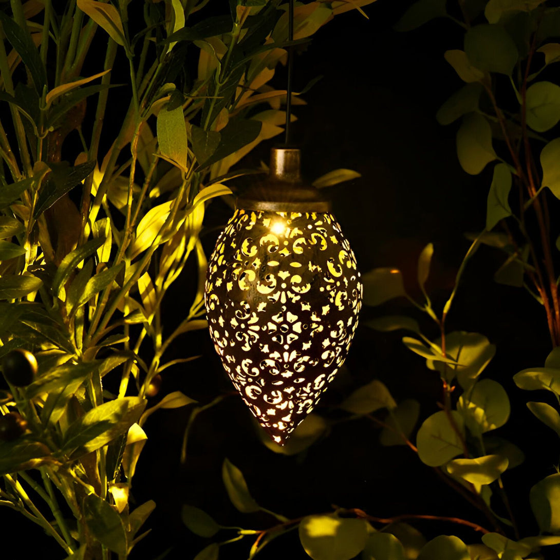 Une lanterne solaire d'extérieur en métal de couleur bronze avec un motif floral ajouré qui diffuse une lumière chaude. La lanterne est suspendue à une branche d'arbre dans un jardin. SU-0023998237,