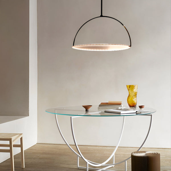 Une table ronde en verre avec des pieds blancs et un plateau en verre transparent. Sur la table se trouve un vase jaune, un bol en bois et des livres. Au-dessus de la table, une lampe noire en forme de demi-cercle est suspendue. CO-2700004558,CO-8923300365,