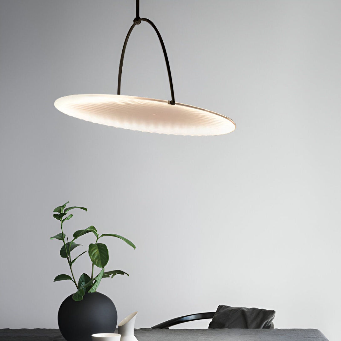 Une suspension design en verre soufflé de forme ronde et plate est accrochée au plafond. Elle est de couleur blanche et diffuse une lumière douce et tamisée. En dessous, une table en bois avec un vase noir contenant une plante verte. CO-2700004558,CO-8923300365,
