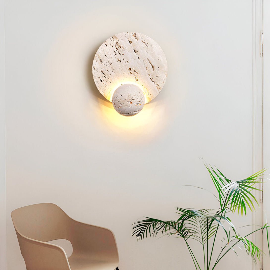Une applique murale ronde en pierre beige avec une lumière chaude. À côté, se trouve un fauteuil beige et un palmier. PI-3050710888,