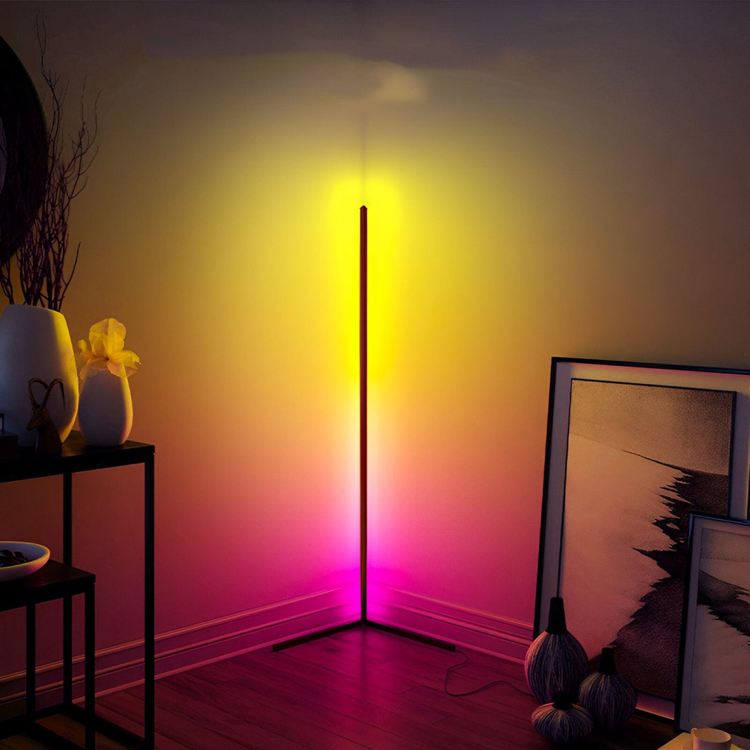L'image montre un lampadaire d'angle à LED RGB qui est placé dans le coin d'une pièce. Le lampadaire est de couleur noire et émet une lumière colorée. La lumière est de couleur rose et jaune. Le lampadaire est placé à côté d'une table sur laquelle se trouve un vase avec des fleurs. Il y a aussi des cadres accrochés au mur. NE-6007763056,NE-5623274994,NE-2525524209,NE-938666142X,