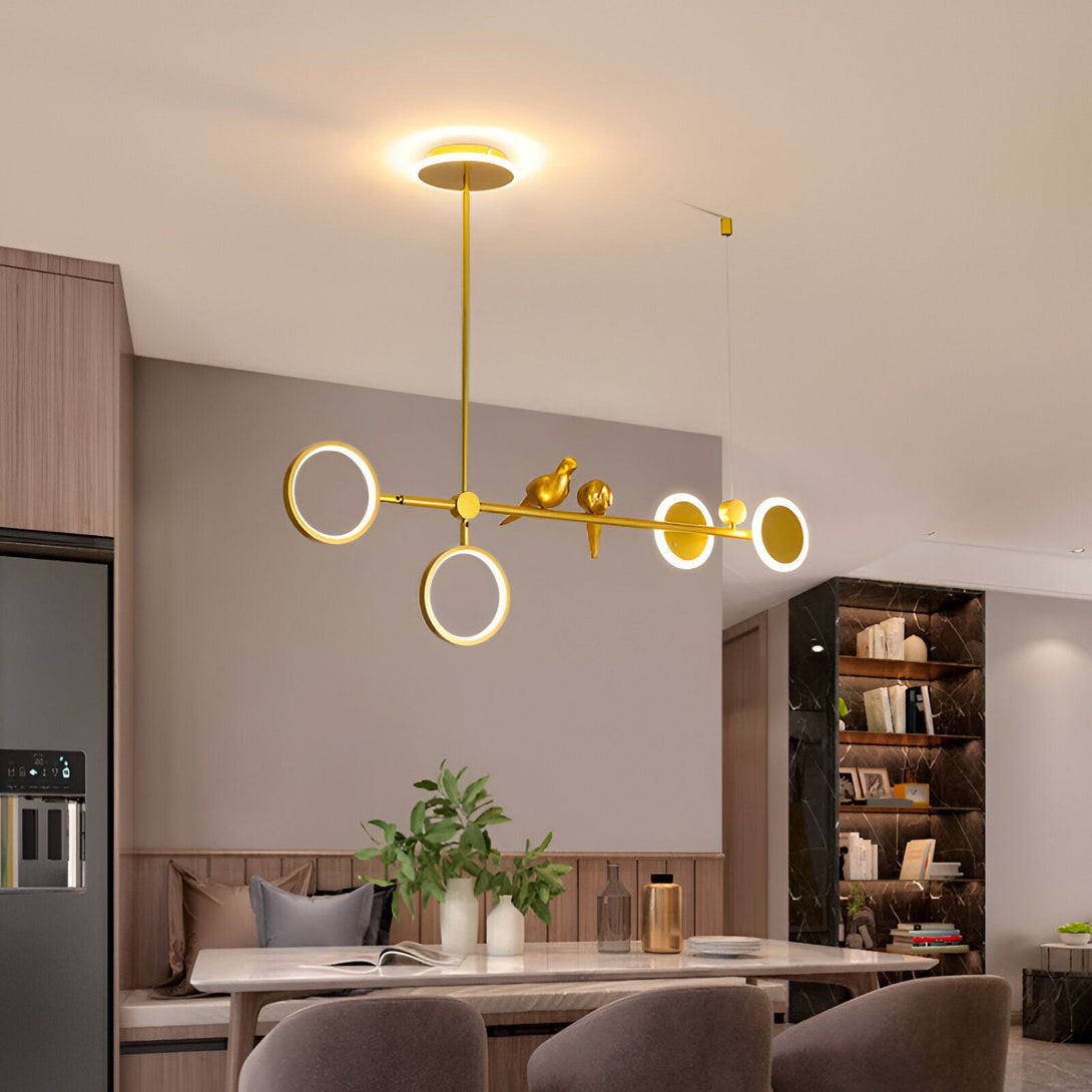 Une image d'un lustre doré avec des oiseaux. Le lustre est composé de plusieurs anneaux de différentes tailles et est suspendu au plafond par un fil doré. Les oiseaux sont perchés sur les anneaux et sont également dorés. Le lustre est allumé et diffuse une lumière chaude et tamisée. OS-708742372X,OS-509121187X,