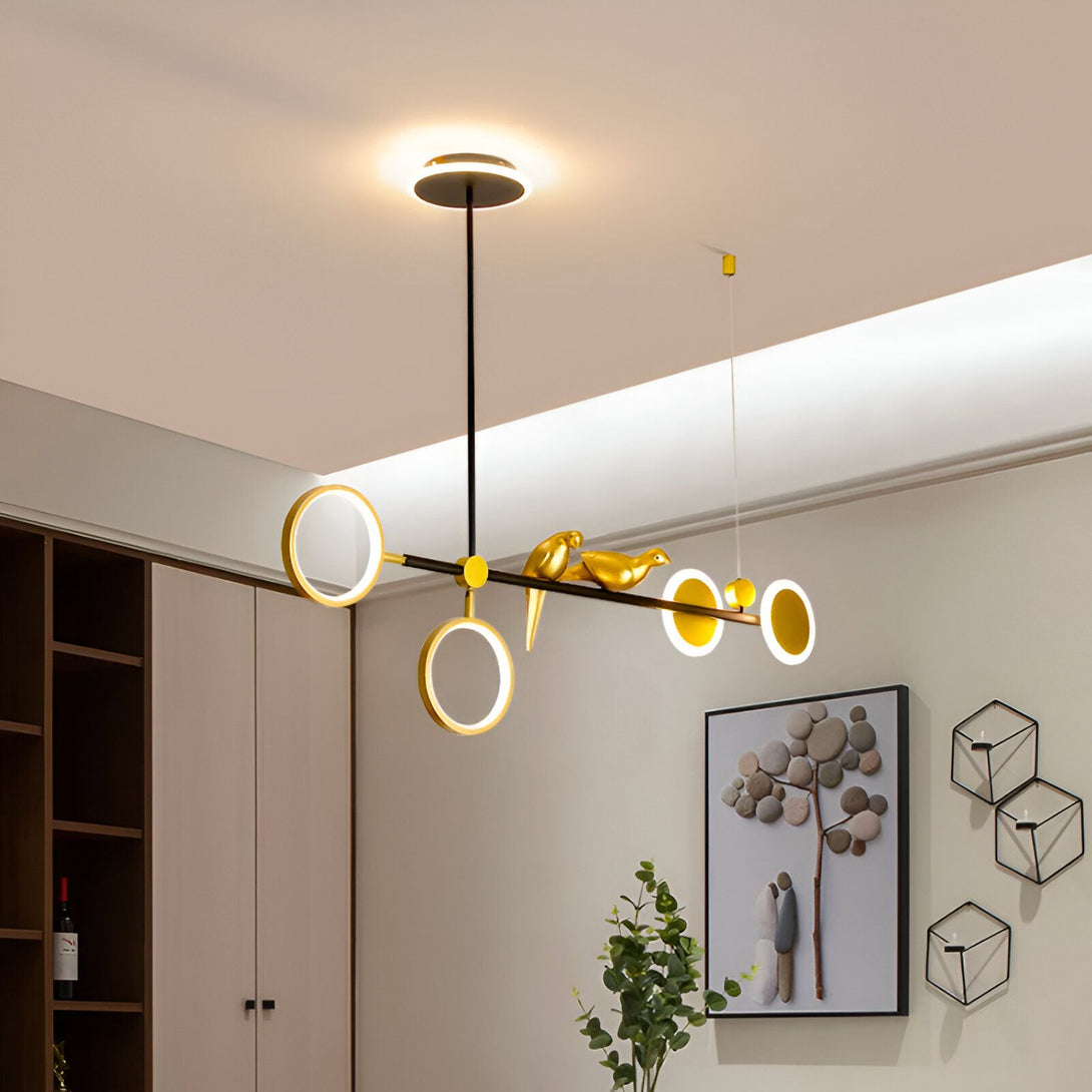 Une image d'un lustre doré avec des oiseaux. Le lustre est composé de plusieurs anneaux de différentes tailles et est suspendu au plafond par un fil noir. Les oiseaux sont perchés sur les anneaux et sont également dorés. OS-708742372X,OS-509121187X,