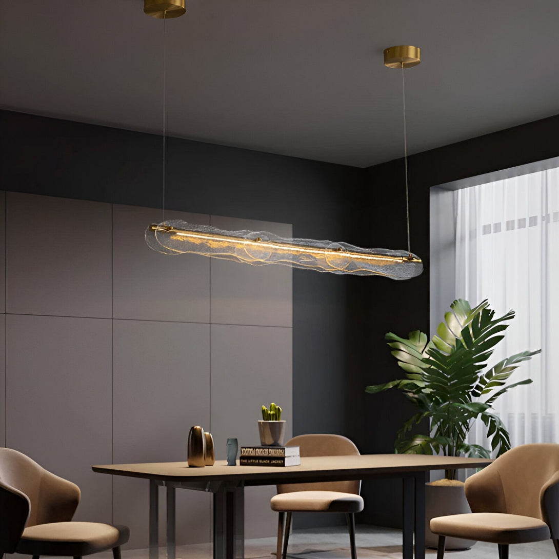 Une vue d'une salle à manger avec une table en bois, des chaises et un lampadaire en verre design suspendu au plafond. Le lampadaire est de forme rectangulaire, avec une structure en métal doré et un diffuseur en verre transparent. Il est suspendu par deux câbles fins. La lumière est allumée et diffuse une lumière douce et tamisée. VE-152859987X,VE-1786463202,VE-6110644315,