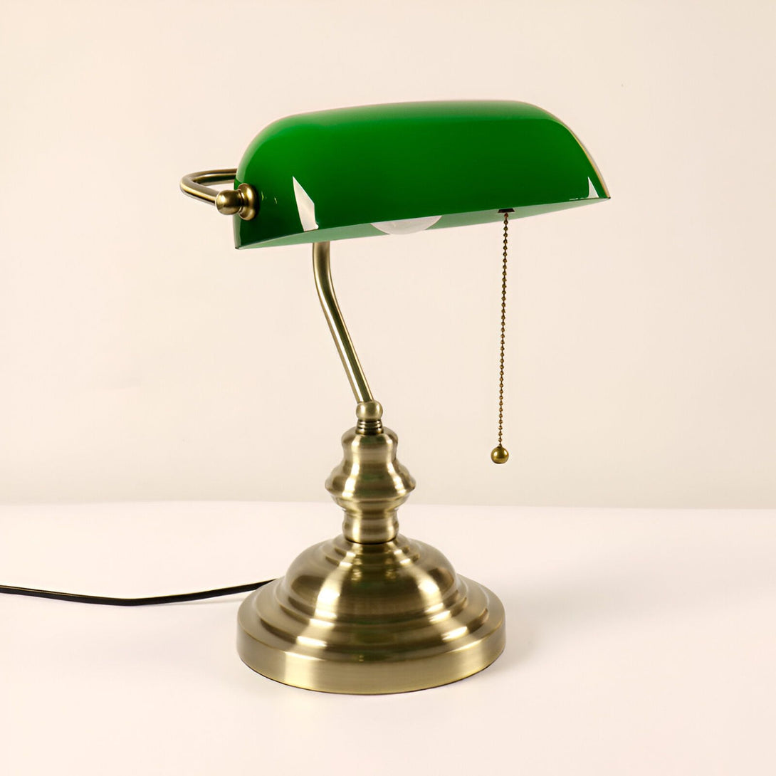 Une lampe de bureau vintage avec un abat-jour vert en verre et une base en métal couleur laiton. La lampe est posée sur une surface blanche. VI-6000882076,VI-6355337148,VI-411590576X,VI-8627305870,