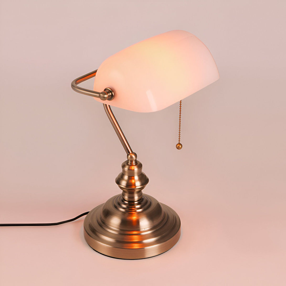 Une lampe de bureau vintage avec un abat-jour en verre blanc et une base en métal couleur bronze. La lampe est allumée et diffuse une lumière chaude. VI-6000882076,VI-6355337148,VI-411590576X,VI-8627305870,