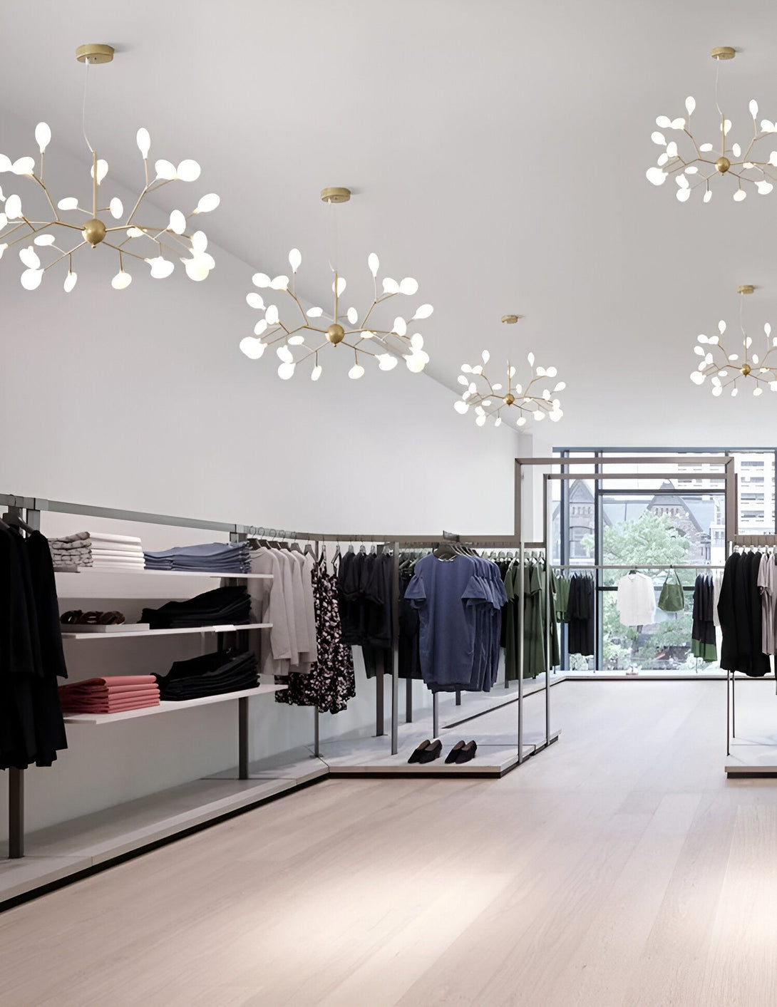 Une image d'un magasin de vêtements avec des vêtements sur des étagères et des portants. Il y a des lumières en forme de branches d'arbre avec des feuilles blanches au plafond. Le sol est en bois et il y a une grande fenêtre à l'arrière du magasin. LU-4202044746,LU-2115327187,LU-2510384147,LU-607350294X,