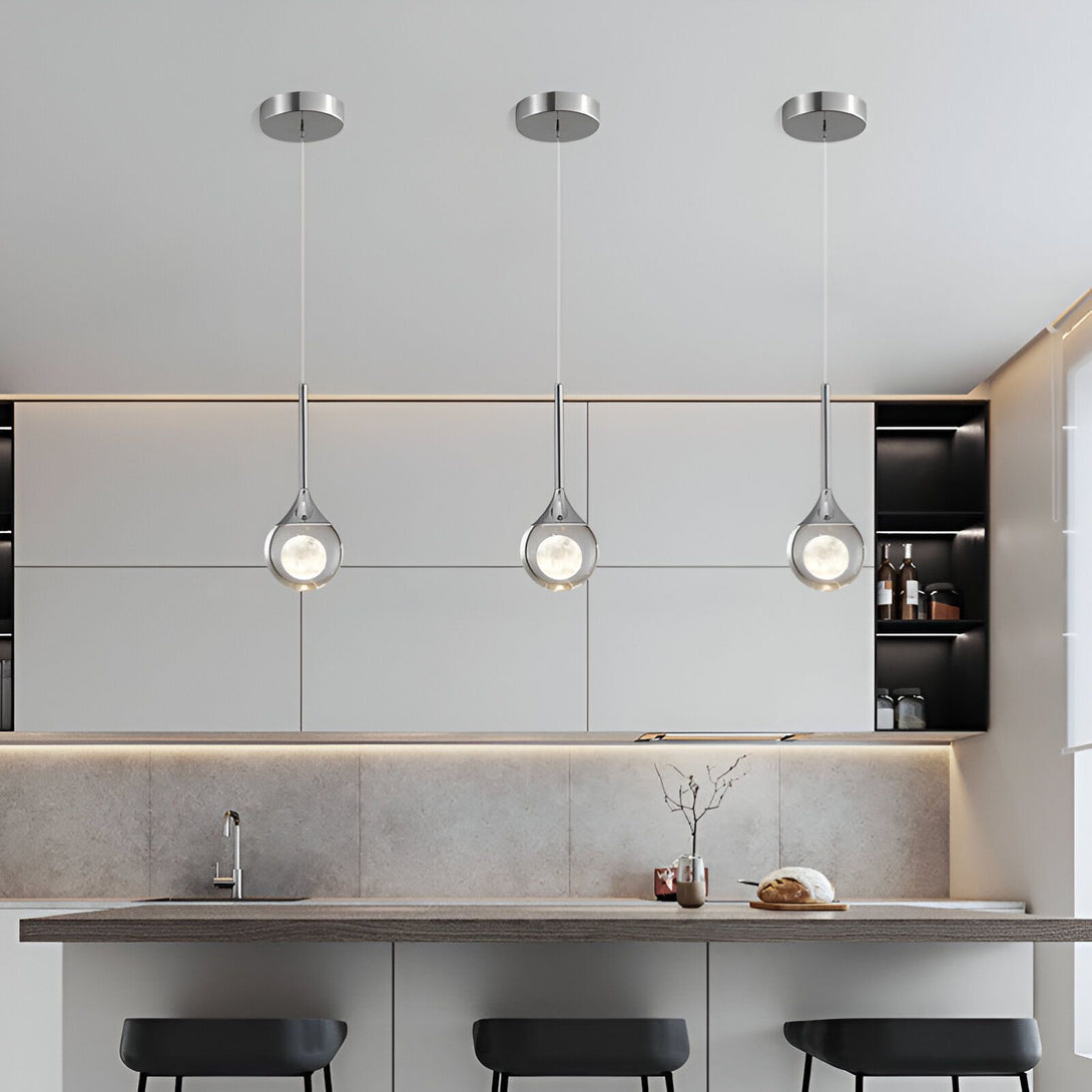 Trois suspensions en forme de goutte d'eau en verre transparent avec une ampoule LED à l'intérieur sont suspendues au-dessus d'un comptoir de cuisine. Les suspensions sont fixées à un plafond blanc. Le comptoir de cuisine est en bois clair et il y a trois chaises noires avec des pieds en métal. IN-6570546732,IN-0202242234,IN-9460160360,IN-5087841252,IN-4331503488,IN-9491831844,