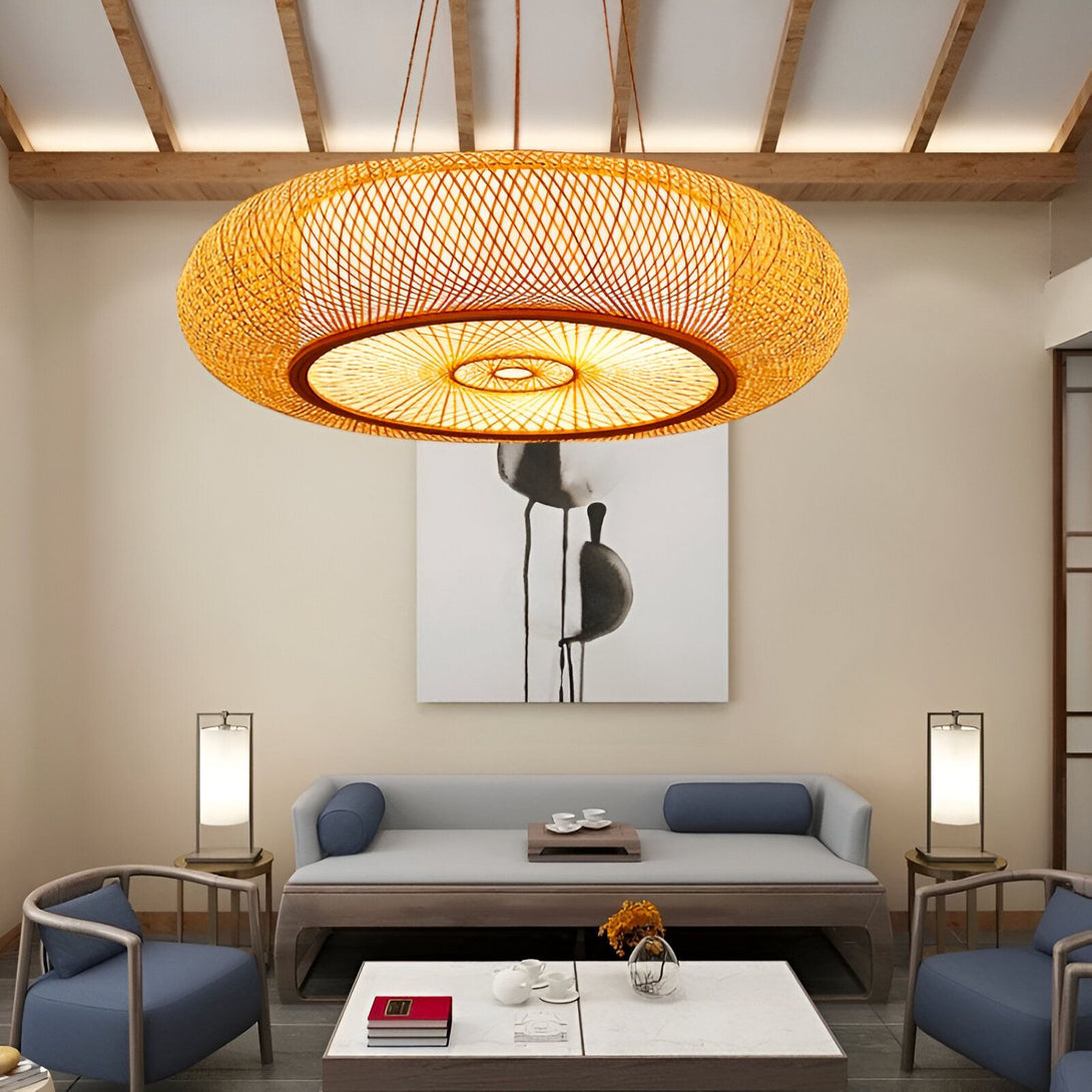 Une lampe en bambou tissé suspendue au plafond d'un salon. La lampe est ronde et ajourée, ce qui permet de diffuser une lumière douce et tamisée. Le salon est meublé d'un canapé, de deux fauteuils et d'une table basse. Les murs sont décorés d'un tableau et de deux appliques. BO-0022616470,BO-2931335754,BO-5250331424,BO-6497583912,