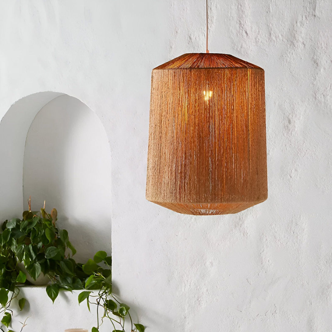 Une lampe suspendue en forme de cage en osier tressé marron clair est accrochée à un fil électrique noir. La lampe est allumée et diffuse une lumière chaude. En arrière-plan, on aperçoit un mur blanc et une plante verte. AD-6453961679,AD-6657275196,AD-8746500626,AD-6764077002,AD-2257925033,AD-5434952707,