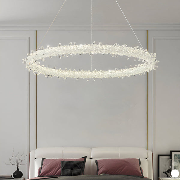 Une image d'un lustre en cristal suspendu au plafond. Le lustre est de forme ovale et est composé de plusieurs branches de cristal qui s'étendent vers l'extérieur. Il est suspendu par des câbles métalliques fins et est éclairé par des ampoules LED. Le lustre est placé au-dessus d'un lit king-size avec une tête de lit capitonnée rose. Il y a deux tables de chevet de chaque côté du lit, chacune avec une lampe dessus. Le sol est recouvert d'un tapis à poils longs blanc.