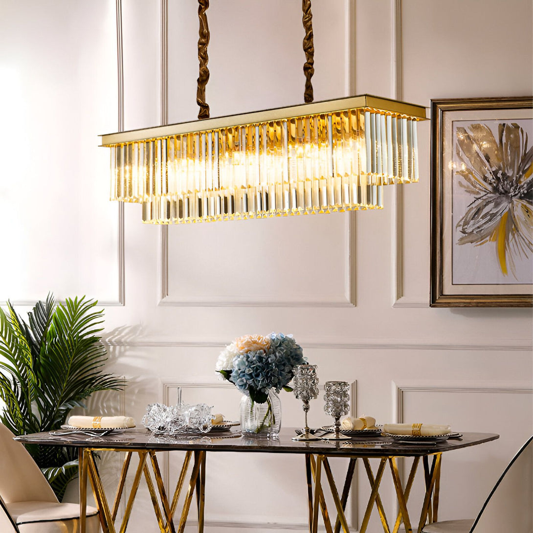 Une image d'un lustre en cristal doré suspendu au-dessus d'une table à manger. Il y a des fleurs sur la table et des chaises autour. Le lustre est rectangulaire et composé de plusieurs rangées de cristaux transparents. Il est suspendu à deux câbles torsadés. La table est en marbre blanc et les chaises sont en bois. Le sol est en parquet. Il y a un tableau accroché au mur du fond. LO-8531375401,LO-5736785838,LO-1500856088,LO-5736656043,