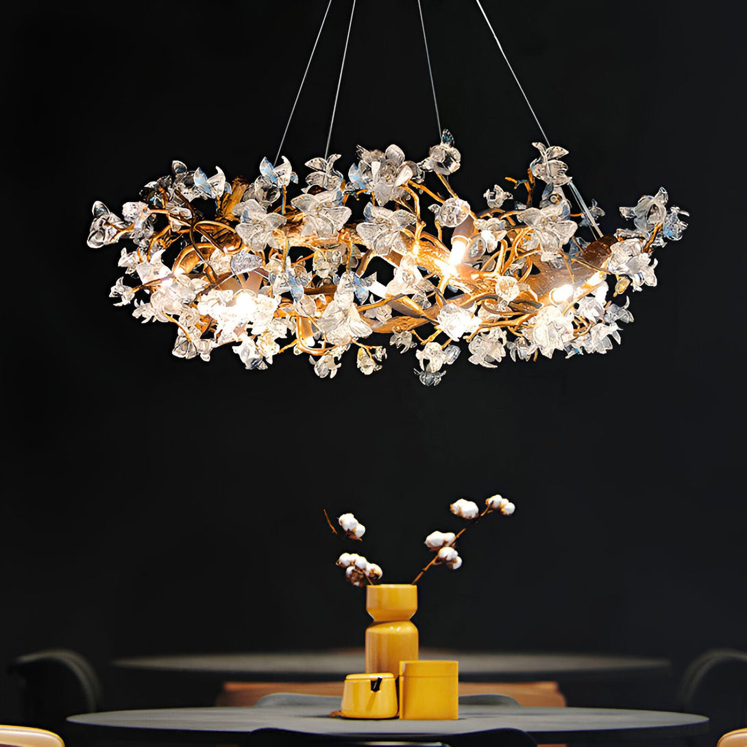 Une image d'un lustre en forme de fleurs en verre soufflé. Il est de couleur or et transparent. Il est suspendu au plafond par des fils métalliques. Il y a une table en dessous avec un vase et des fleurs en coton. Le fond est noir.