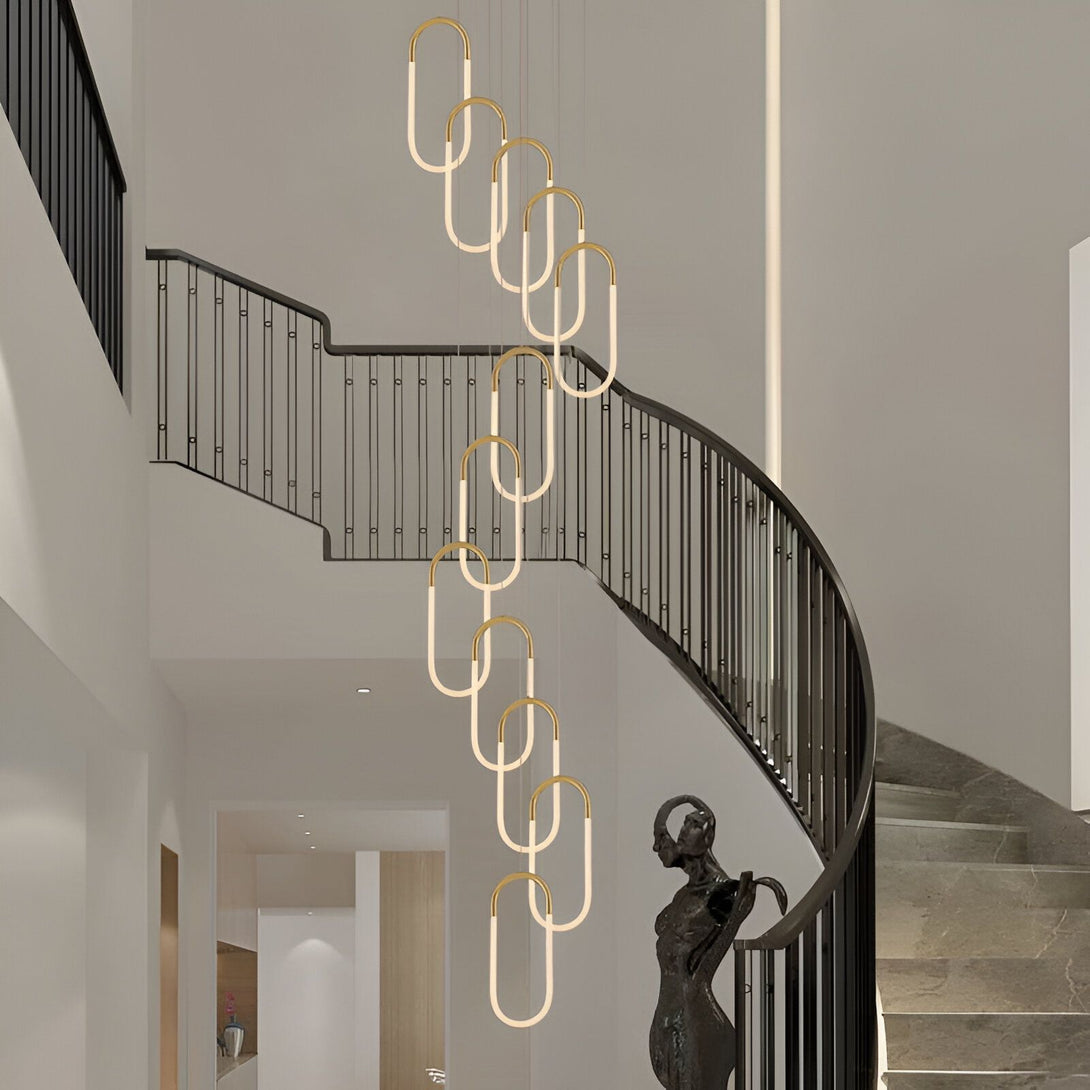 Une image d'un lustre doré suspendu dans une cage d'escalier. Le lustre est composé de plusieurs anneaux ovales en métal doré, et il est éclairé par des lumières LED blanches. La cage d'escalier est en métal noir, et elle est dotée d'une rampe en métal doré. Au sol, on peut apercevoir une statue de femme en bronze. NA-0096577770,NA-4868161806,NA-0573753911,