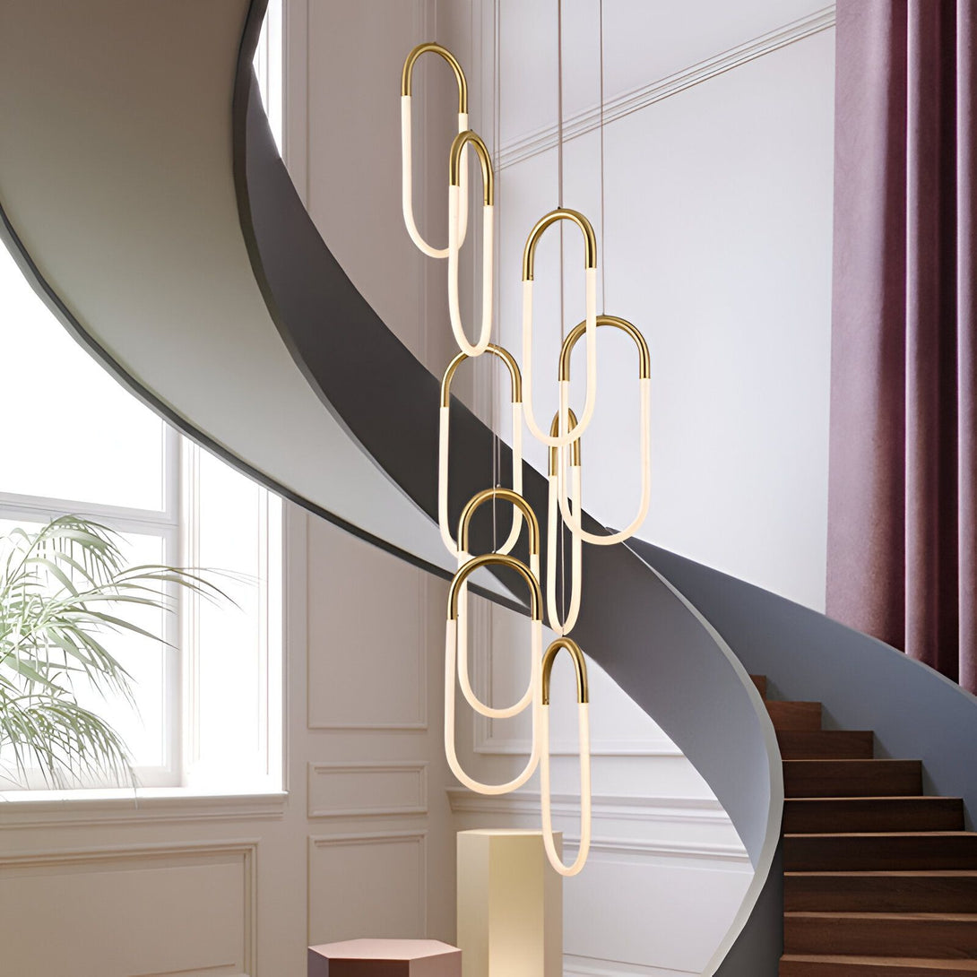 Une image d'un lustre doré suspendu dans une cage d'escalier. Le lustre est composé de plusieurs anneaux ovales en métal doré, qui sont reliés entre eux par des fils métalliques. Les anneaux sont éclairés par des LED blanches, ce qui donne un effet lumineux très élégant. Le lustre est suspendu à un plafond blanc, et il est entouré de murs blancs. Au sol, on peut apercevoir un escalier en bois avec des marches en bois et des contremarches en métal doré. NA-0096577770,NA-4868161806,NA-0573753911,
