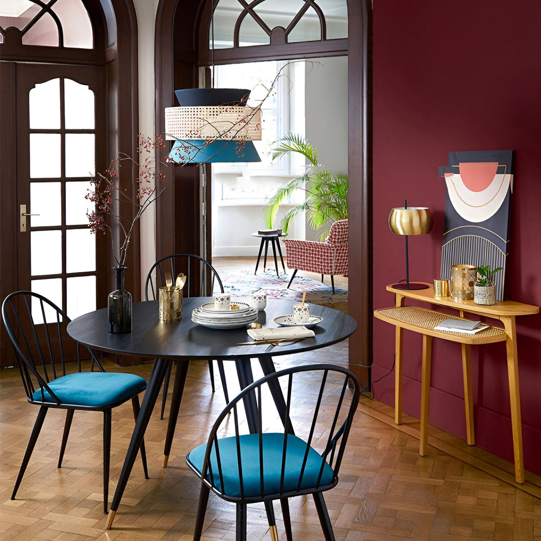 Une table à manger ronde en bois avec quatre chaises rembourrées en velours bleu. Au-dessus de la table, une suspension en osier et métal. À côté de la table, une console en bois avec une lampe dessus. Au fond de la pièce, un grand tapis et un canapé rouge. HE-7629165889,HE-6517411127,HE-800465679X,