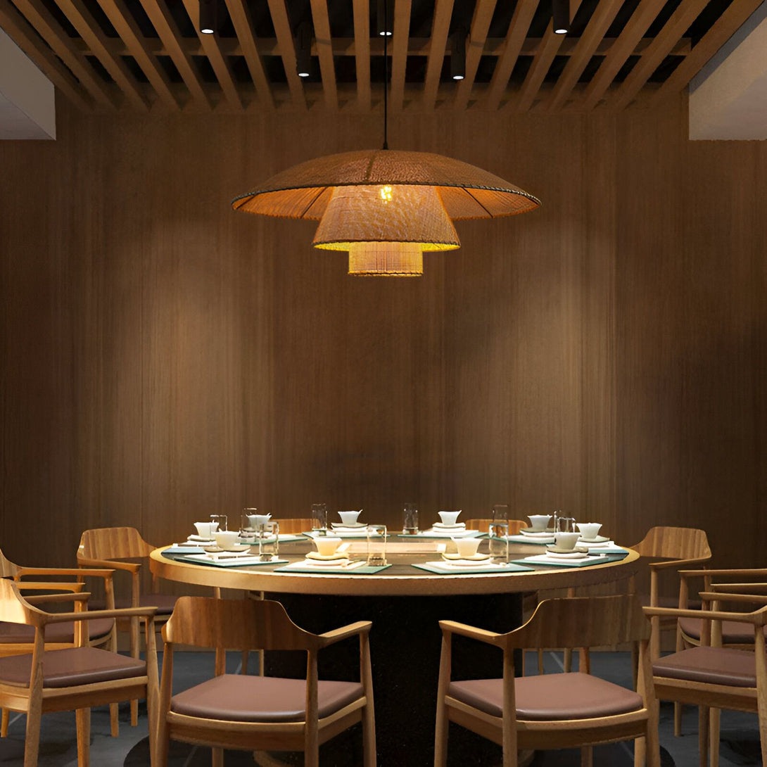 Une grande table ronde en bois est entourée de chaises en bois avec des coussins en cuir marron. Au-dessus de la table, il y a une lampe en osier tressé qui est allumée. Le plafond est en bois et il y a des poutres apparentes. Les murs sont en bois et il y a une porte en bois sur le côté gauche de l'image. JA-9797733807,JA-1062591739,JA-492335965X,JA-1207501026,