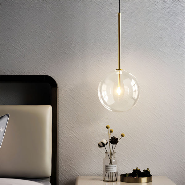 Une image d'une lampe suspendue en verre transparent avec une monture en laiton. La lampe est allumée et émet une lumière chaude. À côté de la lampe, sur une table de chevet, se trouve un vase en verre transparent contenant des fleurs séchées.