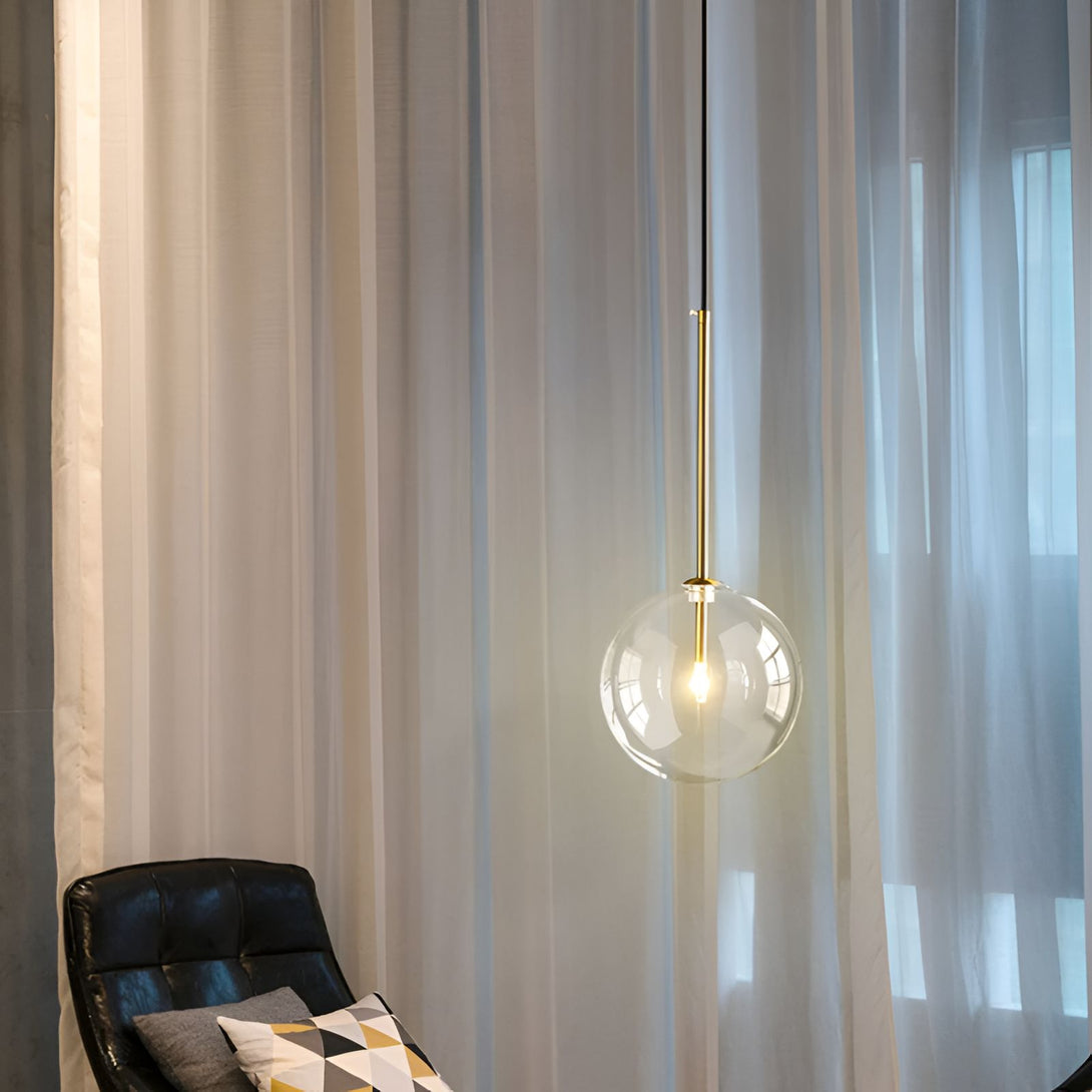Une image d'une lampe suspendue en verre transparent avec une monture en métal doré. La lampe est allumée et émet une lumière chaude. En arrière-plan, on peut voir des rideaux blancs et un fauteuil en cuir noir.