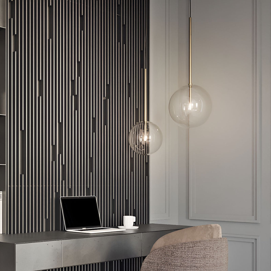 Une image d'une lampe suspendue en verre soufflé avec une finition en laiton. La lampe est suspendue au-dessus d'un bureau en métal avec un ordinateur portable et une tasse de café dessus. Le bureau est placé contre un mur texturé gris foncé.