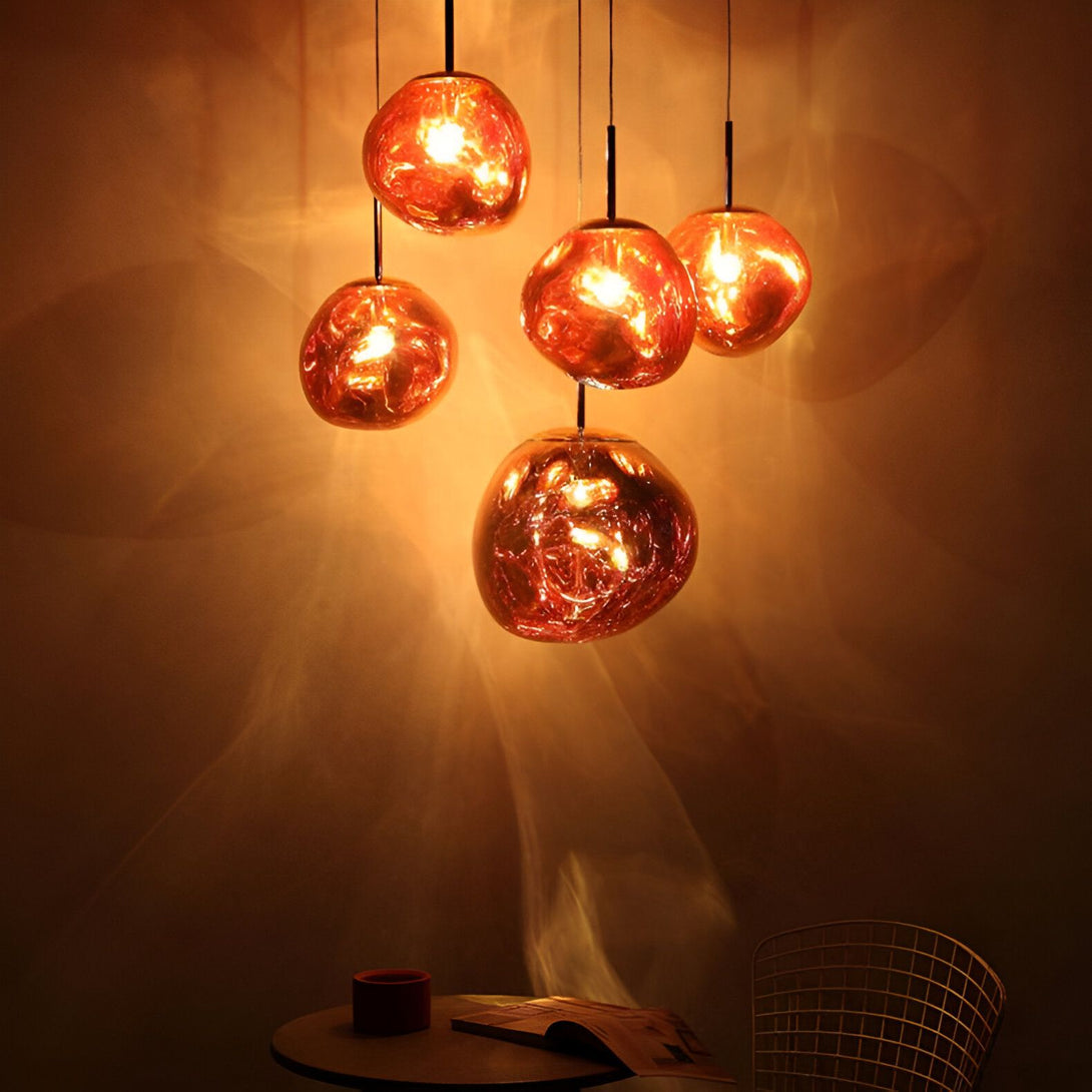 Une image de cinq lampes à suspension en verre soufflé de couleur orange. Les lampes sont de différentes tailles et formes, et elles sont suspendues à des fils noirs. Les lampes sont allumées et émettent une lumière chaude et diffuse. En arrière-plan, on peut voir un mur texturé de couleur marron clair. LA-8821800008,LA-2444977270,LA-6285199337,LA-272961009X,LA-427140456X,LA-929285948X,LA-4420956534,LA-5212907888,LA-5689243493,