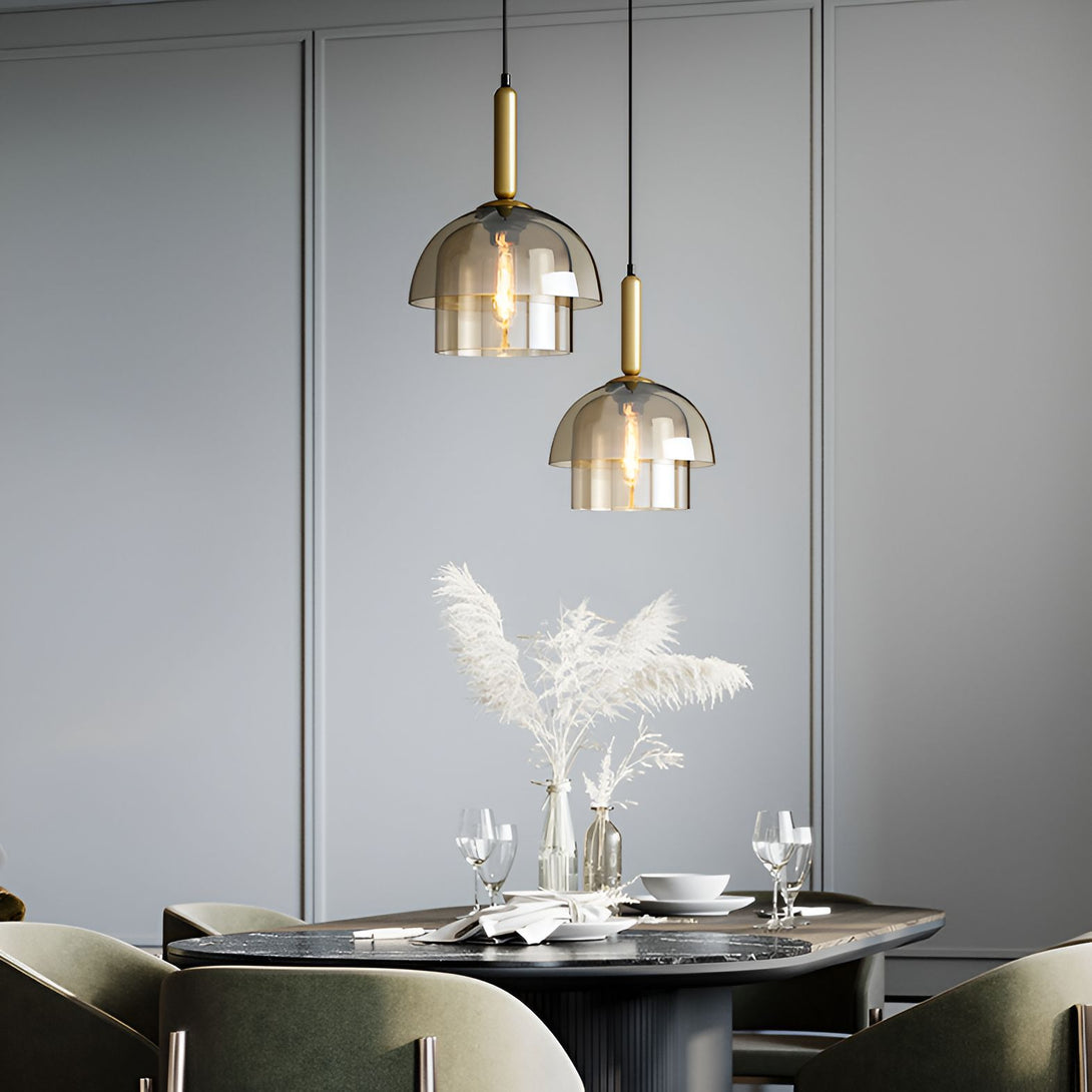 Une image montrant deux suspensions en verre fumé avec des ampoules dorées. Les suspensions sont accrochées au-dessus d'une table à manger en bois avec des chaises vertes. Il y a un vase avec des fleurs séchées sur la table. Le fond est un mur gris avec des moulures. LA-8544977014,LA-4041186528,